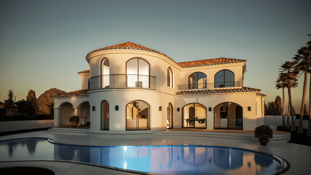 The image shows a Mediterranean-style villa with smooth white stucco walls and terracotta roof tiles. The house features multiple arched windows and doors, with one prominent rounded turret-like section that includes a balcony with wrought iron railings. The villa has a spacious outdoor patio area with steps leading up to it and is surrounded by greenery, including a few palm trees in the background. The overall design is elegant and luxurious, highlighting a blend of traditional Mediterranean and modern architectural elements