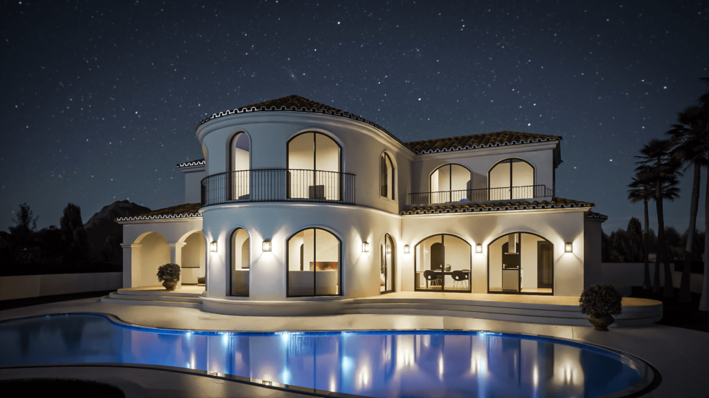 The image shows a Mediterranean-style villa with smooth white stucco walls and terracotta roof tiles. The house features multiple arched windows and doors, with one prominent rounded turret-like section that includes a balcony with wrought iron railings. The villa has a spacious outdoor patio area with steps leading up to it and is surrounded by greenery, including a few palm trees in the background. The overall design is elegant and luxurious, highlighting a blend of traditional Mediterranean and modern architectural elements