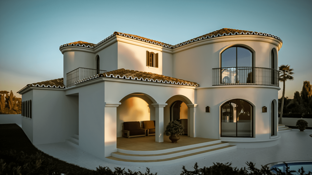 The image shows a Mediterranean-style villa with smooth white stucco walls and terracotta roof tiles. The house features multiple arched windows and doors, with one prominent rounded turret-like section that includes a balcony with wrought iron railings. The villa has a spacious outdoor patio area with steps leading up to it and is surrounded by greenery, including a few palm trees in the background. The overall design is elegant and luxurious, highlighting a blend of traditional Mediterranean and modern architectural elements